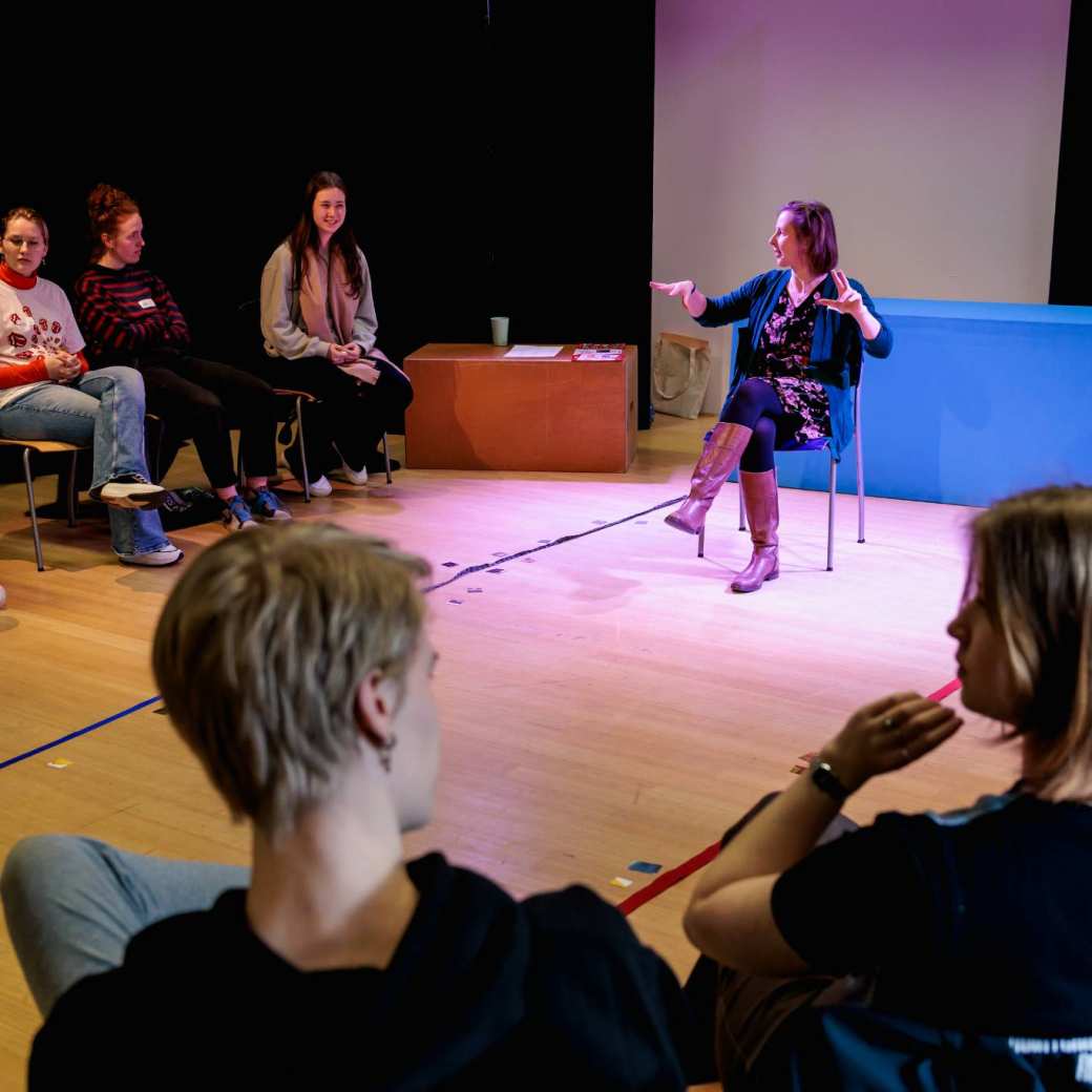 Docent geeft zittend in een kring in een danslokaal uitleg aan deelnemers van het festival 