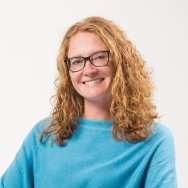 Portretfoto van promovenda Rosa van Doormaal. Zij doet promotieonderzoek bij het project Enactive Mind Autisme. Op de foto heeft ze rood haar, krullen, draagt een bril en zit met blauwe trui in een stoel. 