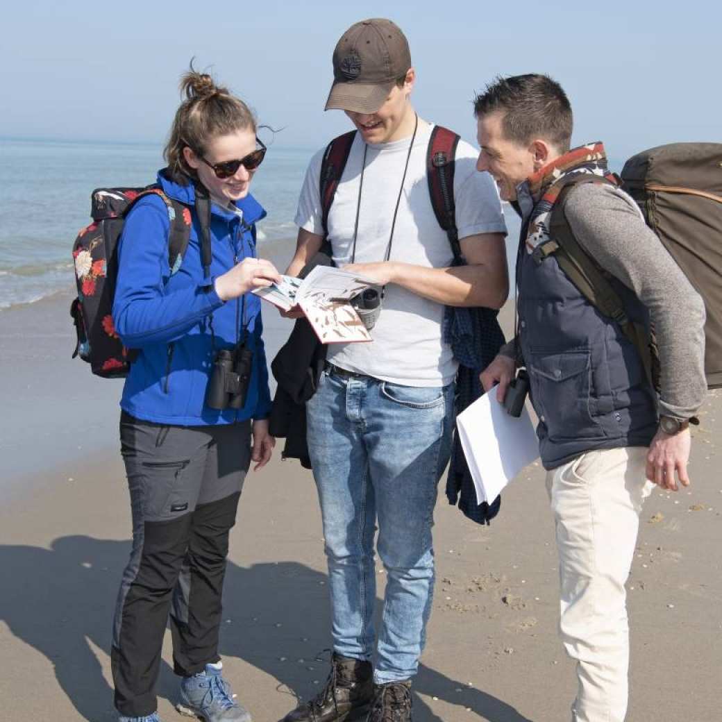 Drie studenten met een rugzak op en een verrekijker in de hand lezen op het strand in een boekje om te kijken waar ze heen moeten.