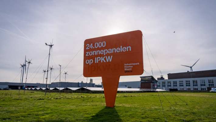 Foto van het Industriepark Kleefse Waard (IPKW) bij de ingang van het park met een bord over zonnepanelen capaciteit van het IPKW en met windmolens op de achtergrond. Academie Engineering en Automotive.