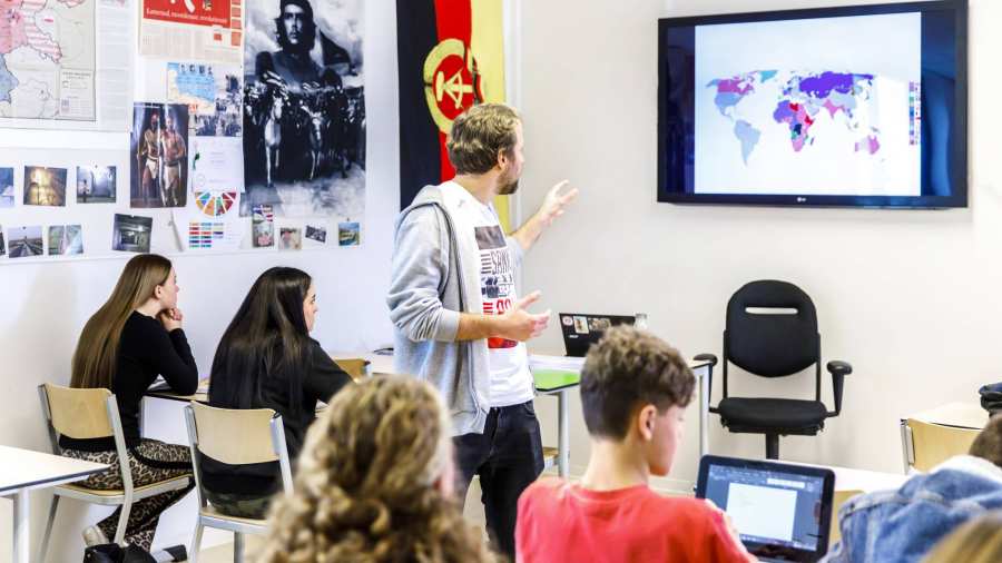 Docent staat midden in de klas en geeft uitleg aan zijn leerlingen die naar het smartbord kijken waar een wereldkaart op staat. 