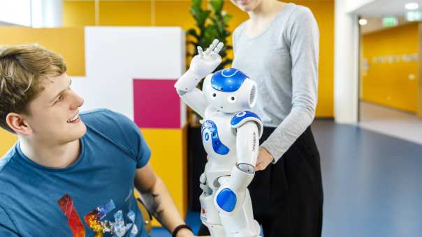 Een mannelijke student International Social Work kijkt naar een zorgrobot voor het vak Sociale Technologie, op de achtergrond staat een vrouwelijke medestudent.