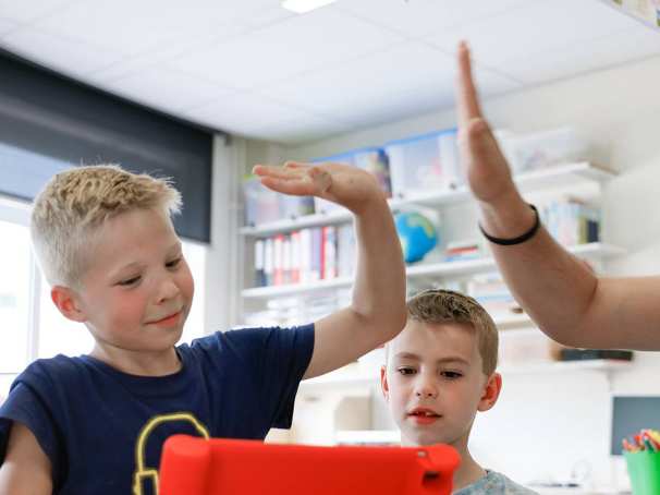 Leraar zit met drie kinderen achter de iPad en geeft Ã©Ã©n van die leerlingen een high-five. 