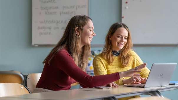 2 studenten kijken lachend en wijzend naar laptop. in een klaslokaal.