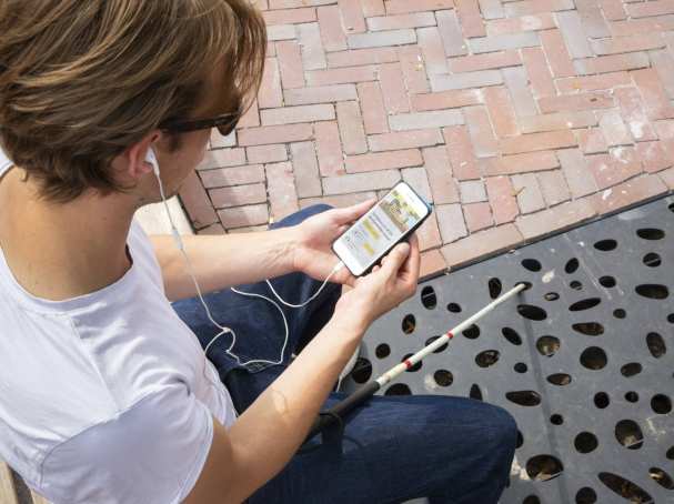 Jongen zit op een bankje en is met zijn mobiele telefoon bezig. Voor hem ligt zijn taststok.