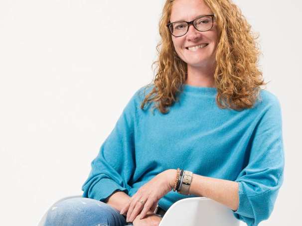 Portretfoto van promovenda Rosa van Doormaal. Zij doet promotieonderzoek bij het project Enactive Mind Autisme. Op de foto heeft ze rood haar, krullen, draagt een bril en zit met blauwe trui in een stoel. 