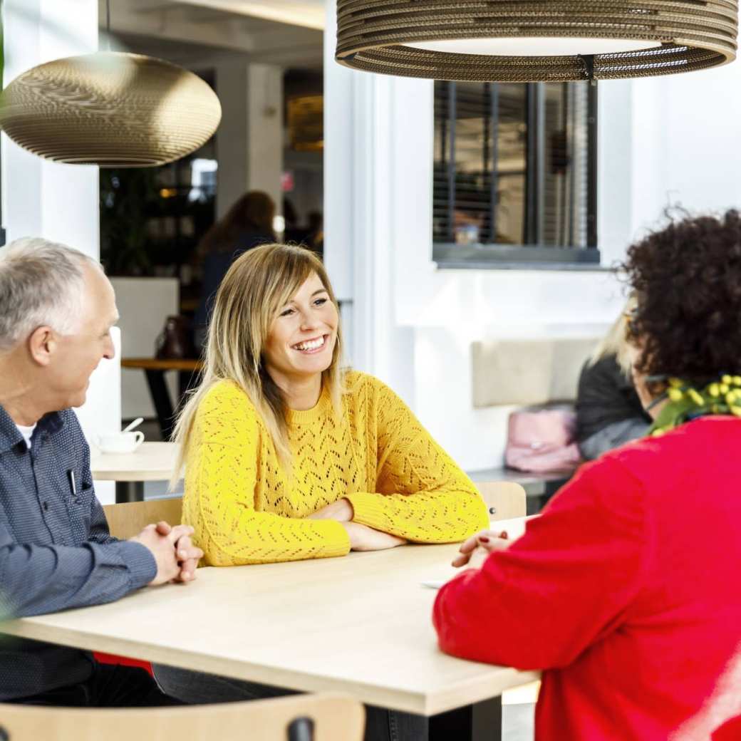 Een man en twee vrouwen zitten pratend aan een tafel. Vrouw met gele trui lacht tijdens het gesprek.