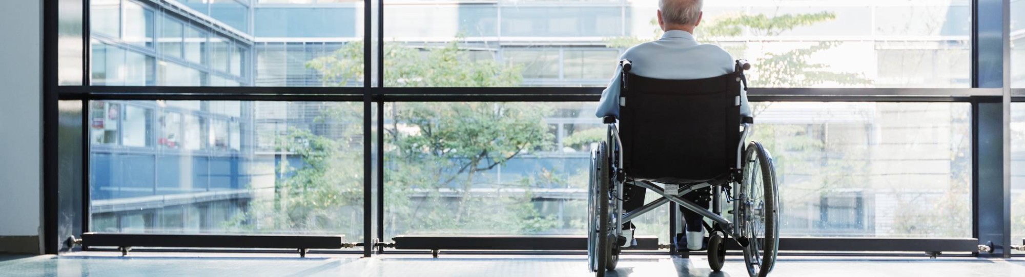 oudere man in rolstoel kijkt uit raam in een het ziekenhuisgang