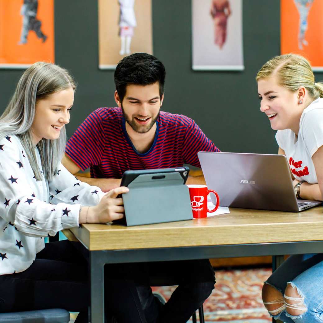 Drie studenten kijken lachend naar de ipad waar een medestudent iets aanwijst.