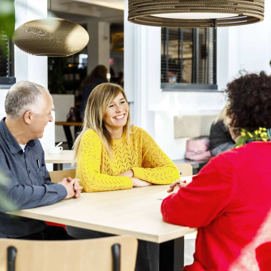Twee vrouwen en een man pratend aan een tafel. Een vrouw met een gele trui lacht tijdens het gesprek