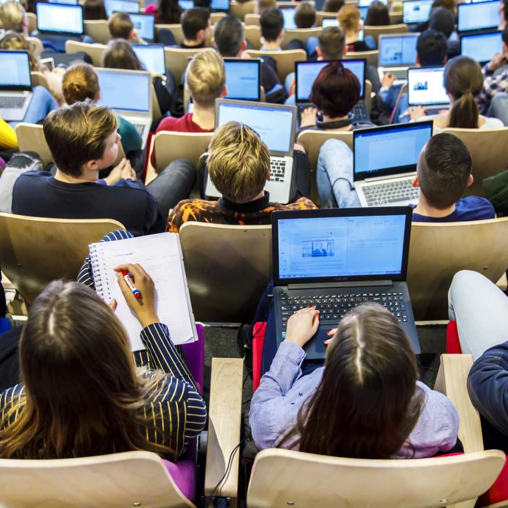 Foto van boven gemaakt van studenten die met de laptops opengeklap in de collegezaal zitten.