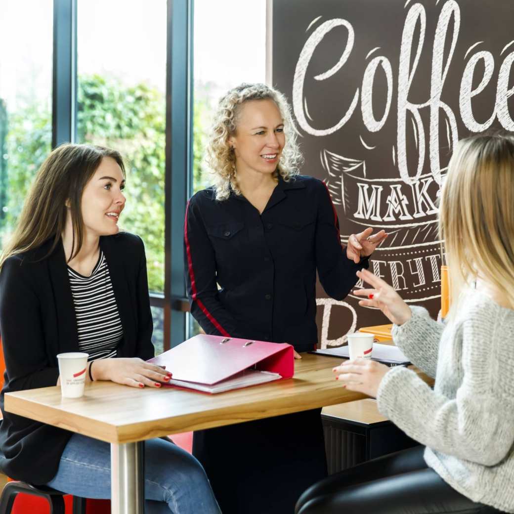 3 vrouwen aan tafel voor een grijze muur met de tekst ''coffee'' bij logistiek duaal