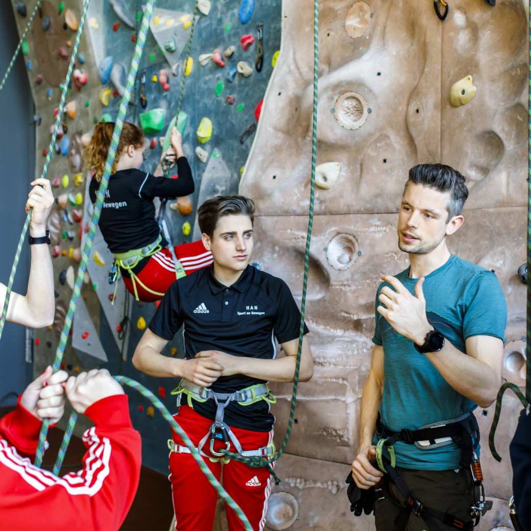 Vier studenten van de HAN krijgen uitleg van hun gymdocent over het beklimmen van de klimwand.