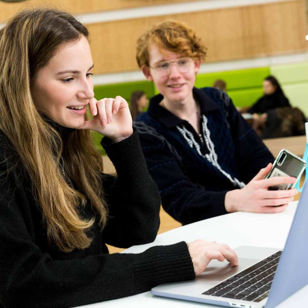 259514 ALPO student met smartphone kijkt mee op de laptop scherm van zijn klasgenoot