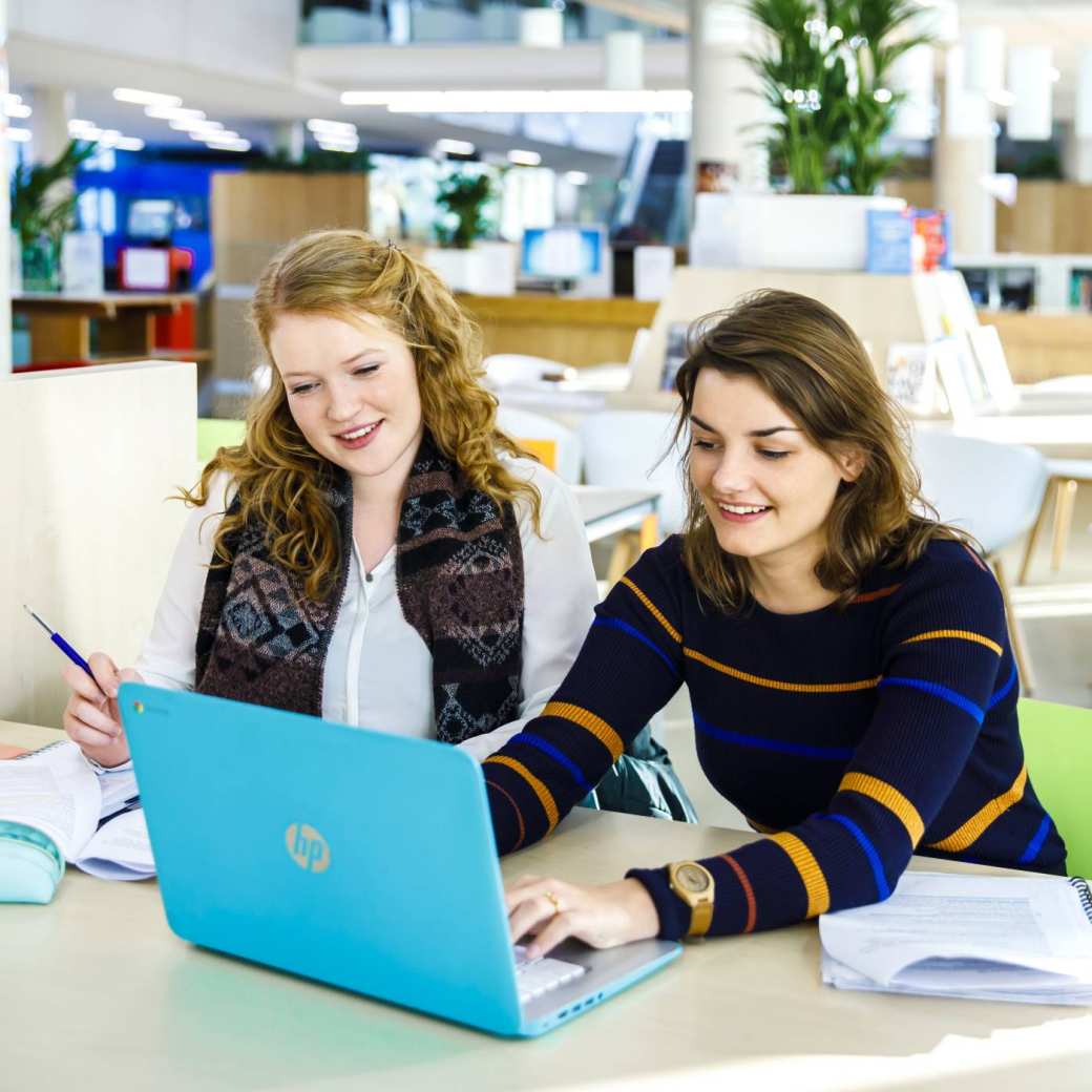 Twee studenten werken samen achter de laptop in het I/O gebouw en kijken lachend naar het scherm.