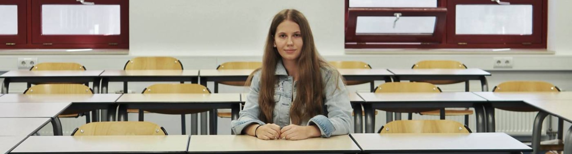 Portretfoto van Annemae de Vries, ze zit in een klaslokaal aan een tafel op een stoel. Haar lange donkerblonde haar zit los, ze draagt een lichtblauw spijkerjasje, witte jeans en witte sneakers
