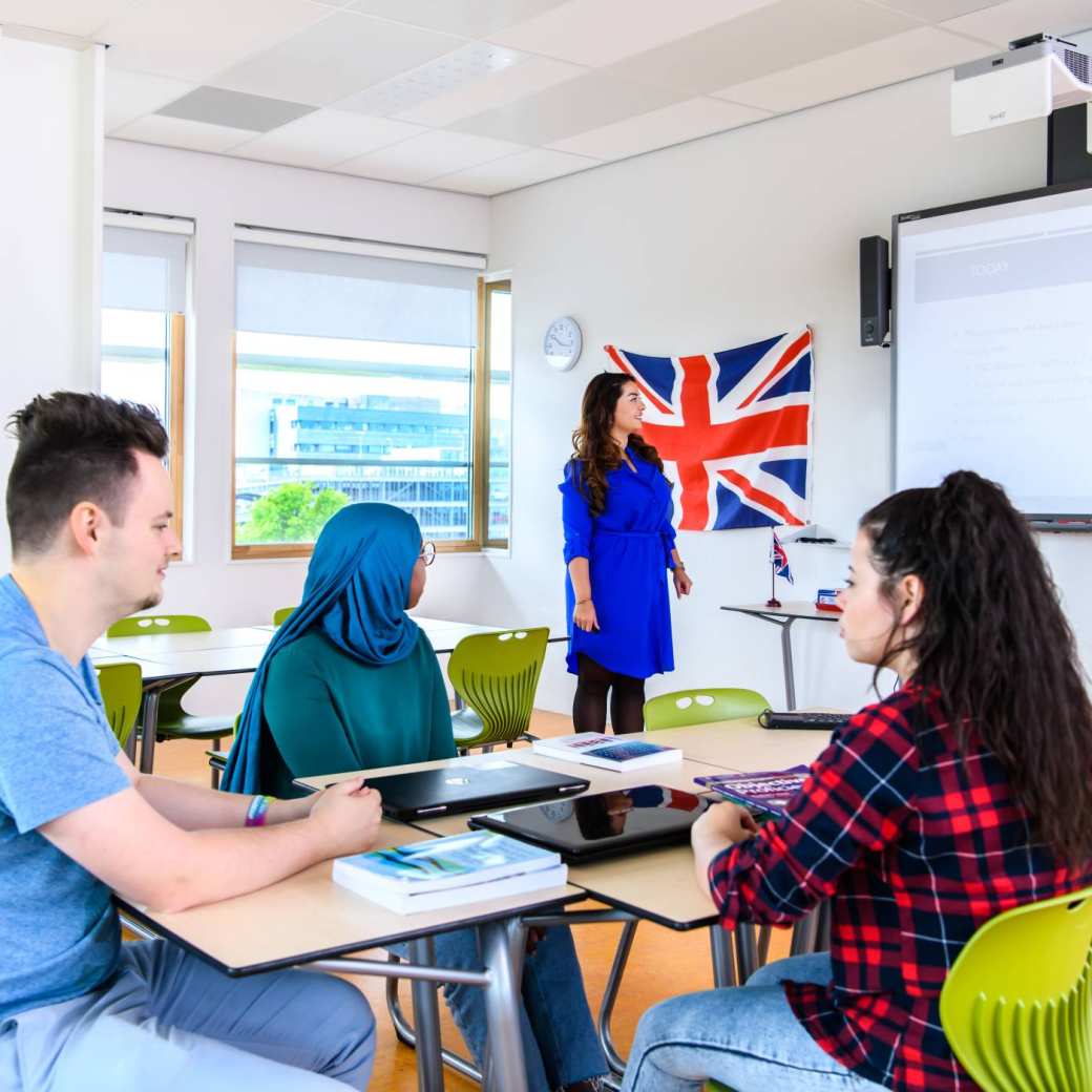 Docent geeft les aan haar drie studenten in een lokaal met de Engelse vlag aan de muur.