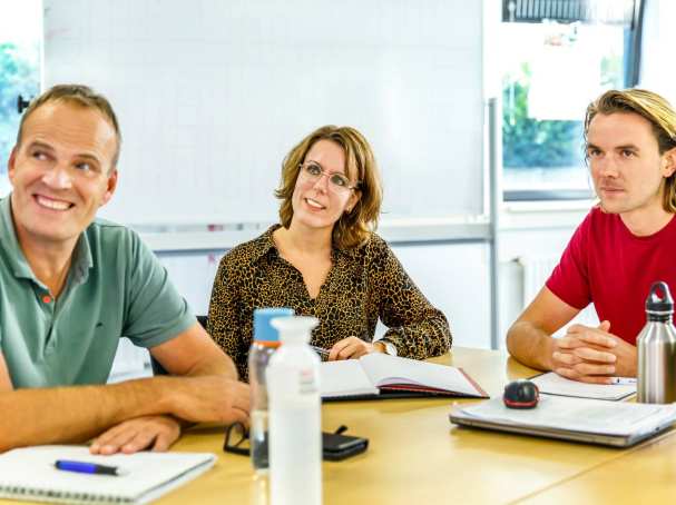 Drie studenten van de master ontwerpen van eigentijds leren kijken lachend aandachtig richting een docent. 