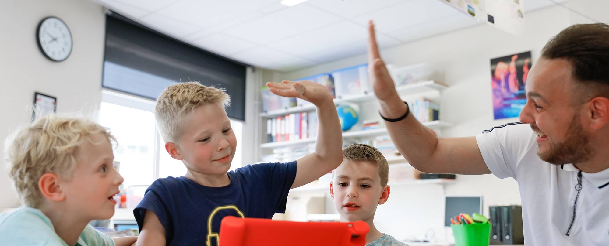 Leraar zit met drie kinderen achter de iPad en geeft één van die leerlingen een high-five.