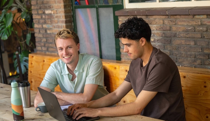 Twee studenten zitten aan tafel te studeren achter de laptop en lacht naar camera