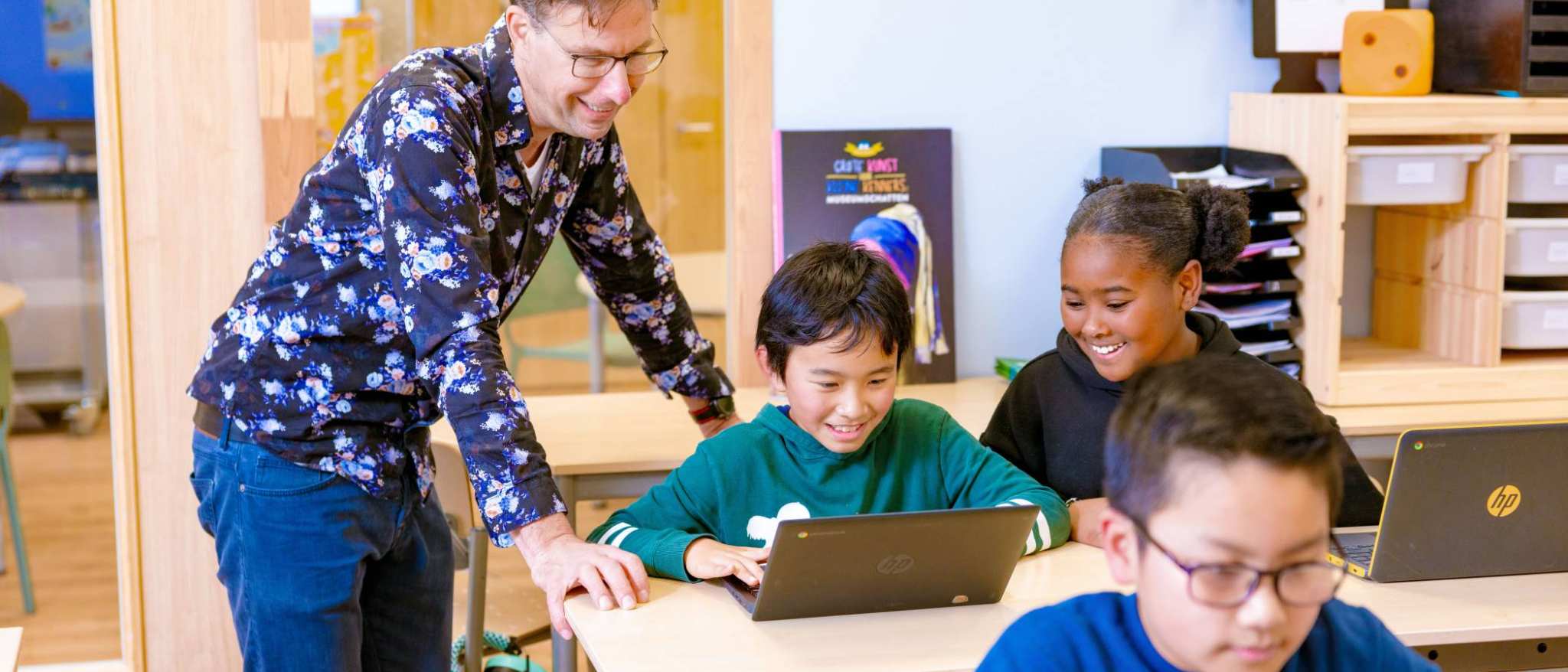 Groep 5,6,7 op de school Da Vince. De leraar en twee leerlingen lachen die achter hun laptop aan het werk zijn.