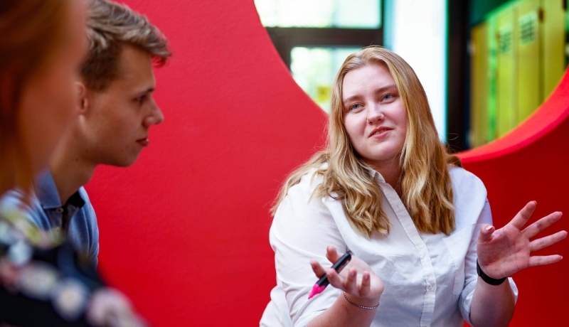 Jonge vrouw met blond haar en witte blouse heeft een marker in haar hand en praat met 2 jongens