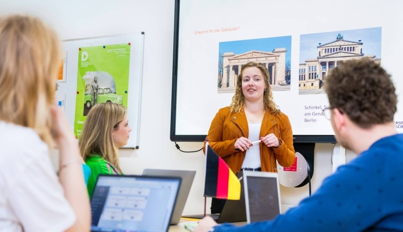 Student leraar Duits in oranje/bruin jasje geeft een lesje aan haar medestudenten. Ze staat voor een PowerPoint.
