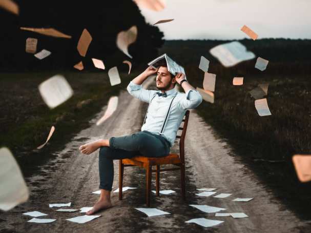 man met wit overhemd, vlinderdas en bretels zit op een stoel, midden op een zandweg. Hi houdt een boek boven zijn hoofd en losse pagina's vliegen in het rond