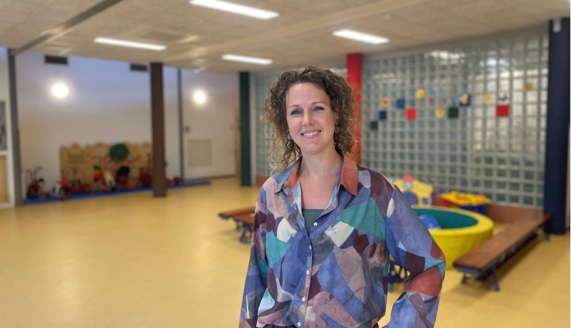 Marjolein Ubbink heeft donkerblonde krullen en draagt een blauw/paarse blouse met een blok print. Ze staat met haar hand in de zij en lacht breeduit.