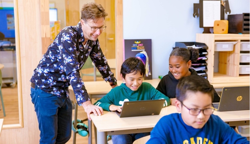 Groep 5,6,7 op de school Da Vince. De leraar en twee leerlingen lachen die achter hun laptop aan het werk zijn. 
