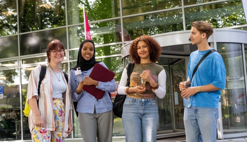 Vier studenten staan bij gebouw LvS10 bij draaideur, buiten. Op een rijtje.