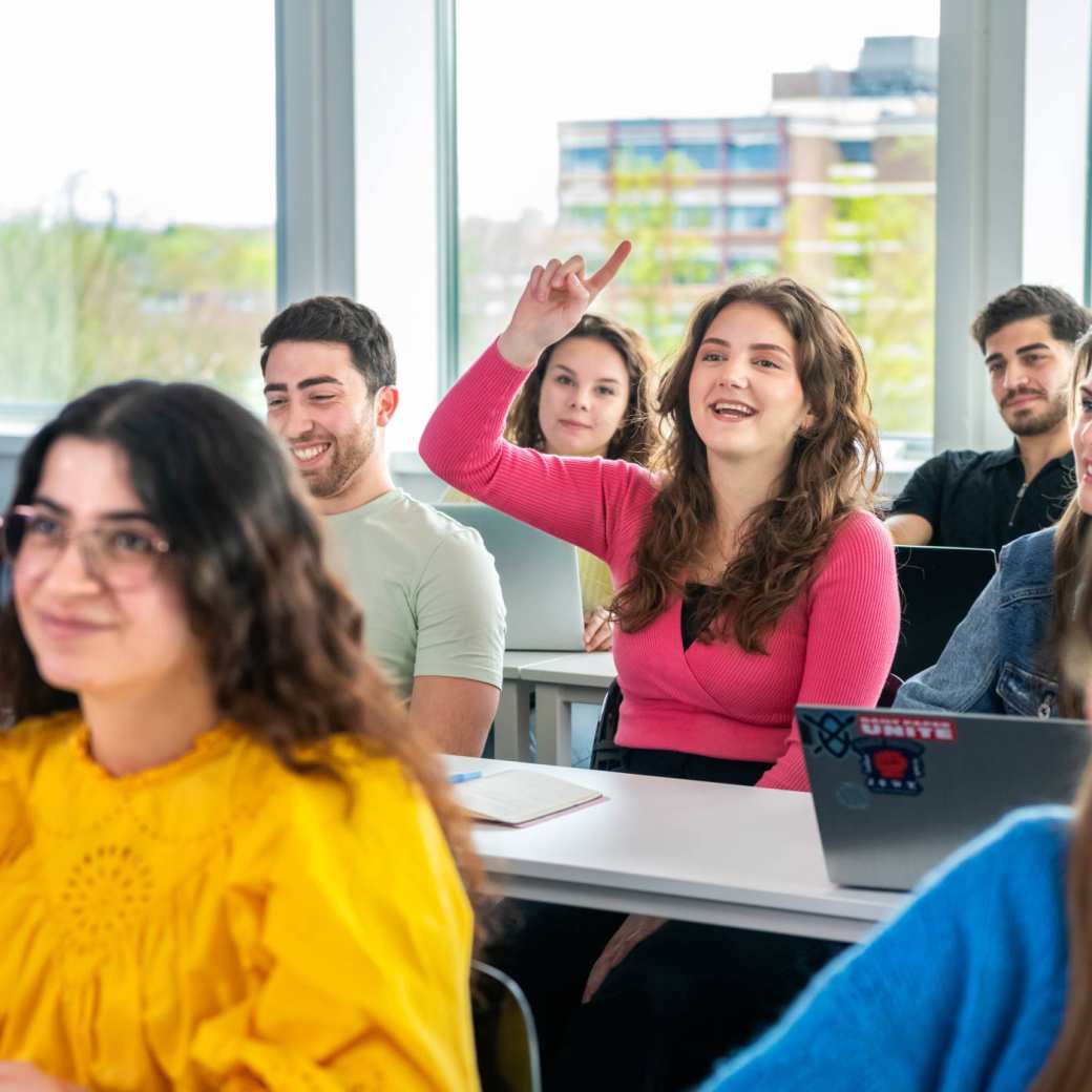 Studenten zitten in de klas, 1 student steekt een vinger omhoog om een vraag te stellen.