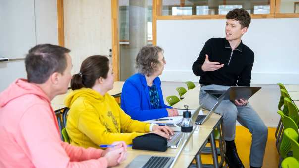 Foto Master Academie Educatie. 4 masterstudenten zitten in een lokaal. 1 van hen zit op een bureau en legt met een laptop in z'n hand wat uit aan de rest.