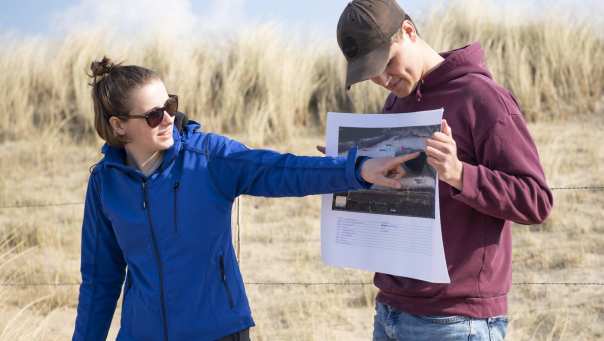 Twee studenten doen veldwerk op het strand. Een student houdt een foto vast en de andere student wijst iets aan op de foto. 