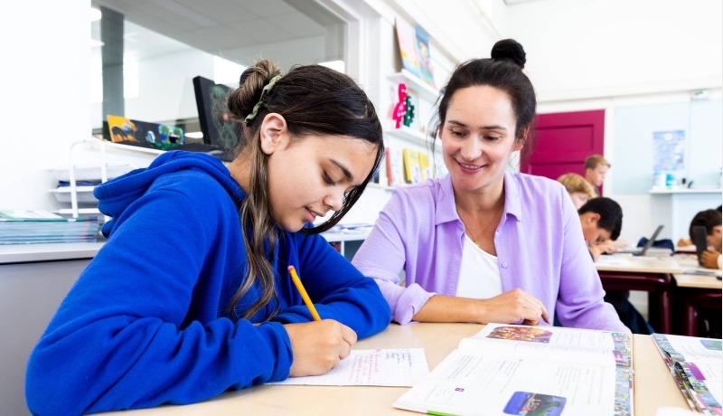 De lerares van de opleiding leraar basisonderwijs geeft persoonlijke uitleg aan leerling aan haar tafel