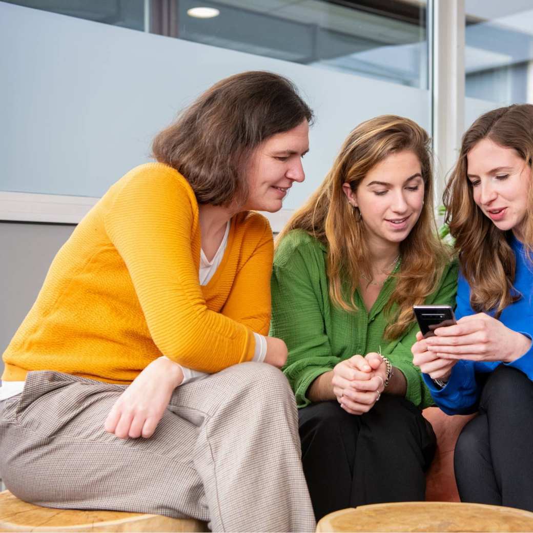 3 Studenten zitten samen op krukjes en kijken op een telefoon.
