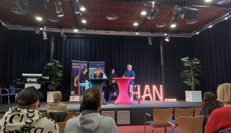 sfeerbeeld van de opening van de week tegen armoede. Mensen zitten op een stoel in een zaal met rode vloerbedekking.