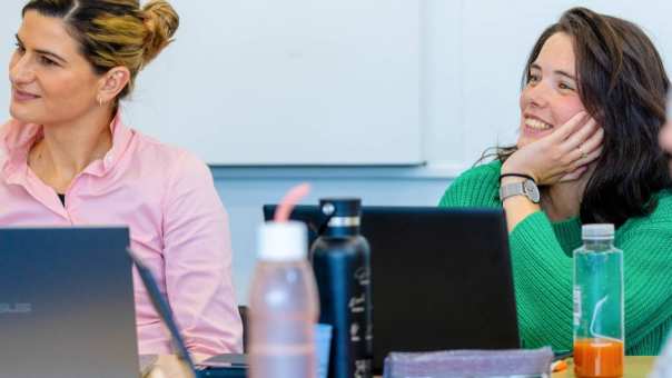 Een vrouw in een groene trui en roze trui zitten in het leslokaal met beide een laptop op tafel