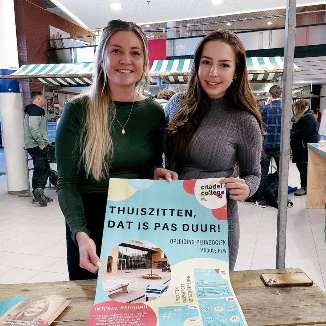 Hanne Wanink heeft blond haar en een groene top en draagt een gouden ketting. Romy Burghouts heeft een grijze jurk aan en donkere haren. Ze houden een poster in hun hand van het project met daarop Thuiszitten, dat is pas duur.