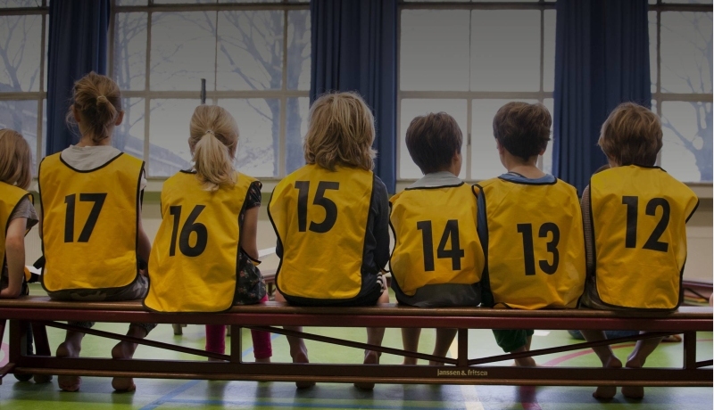 Kinderen in een gymzaal met hesjes aan op de rug gefotografeerd
