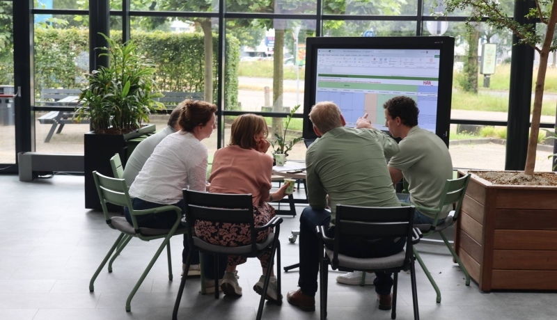 Een subgroepje van de sessie DMA Samen uitvoeren bespreekt de resultaten bij een beeldscherm. Zittend, gebogen over papieren.