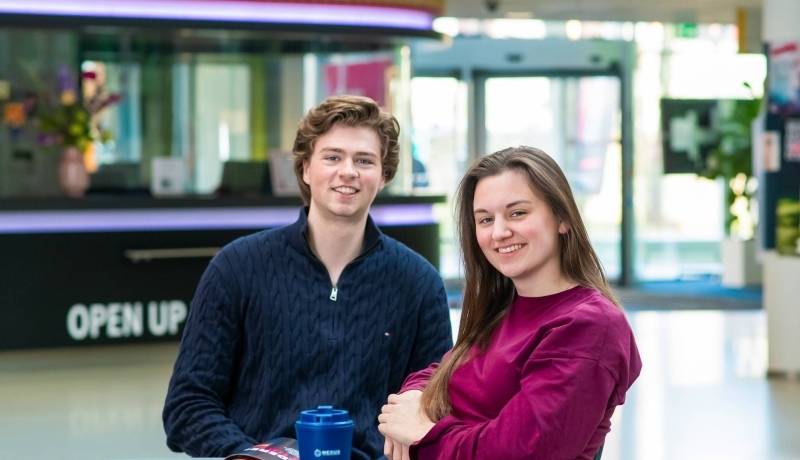 Twee studenten hbo Bedrijfskunde zitten aan een tafel en kijken in de camera