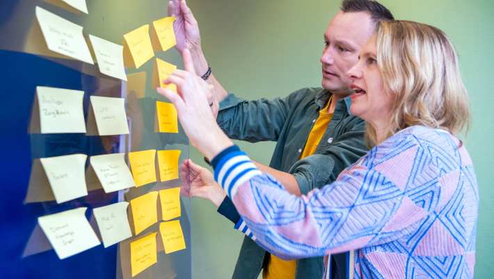 Man en vrouw kijken samen naar een glazen bord met gekleurde post-its erop geplakt.