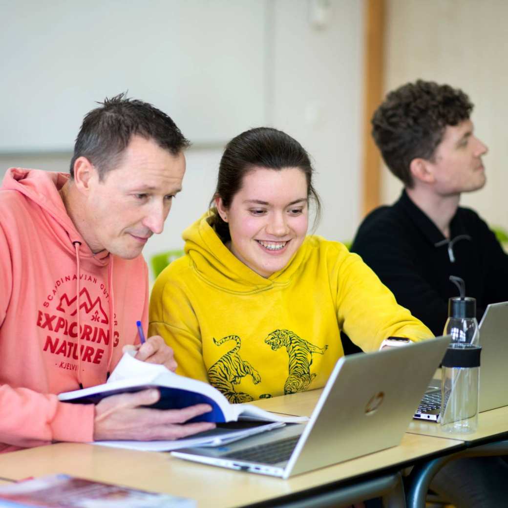Foto Master Academie Educatie. 3 masterstudenten zitten aan hun bureaus. 2 ervan kijken samen op een laptop met een boek in de hand.