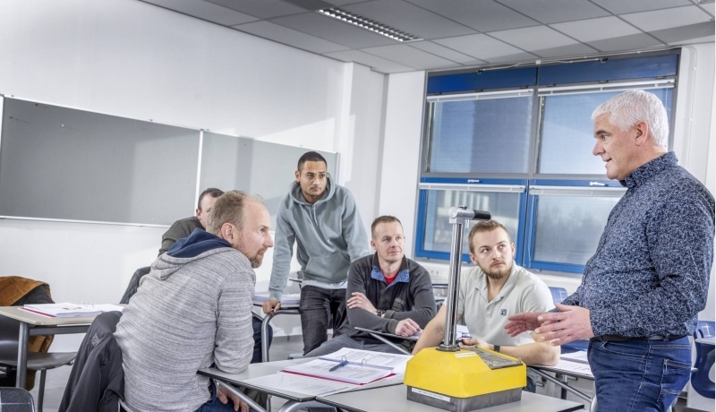 5 mannen zittend en staand aan een tafel luisteren aandachtig naar de docent