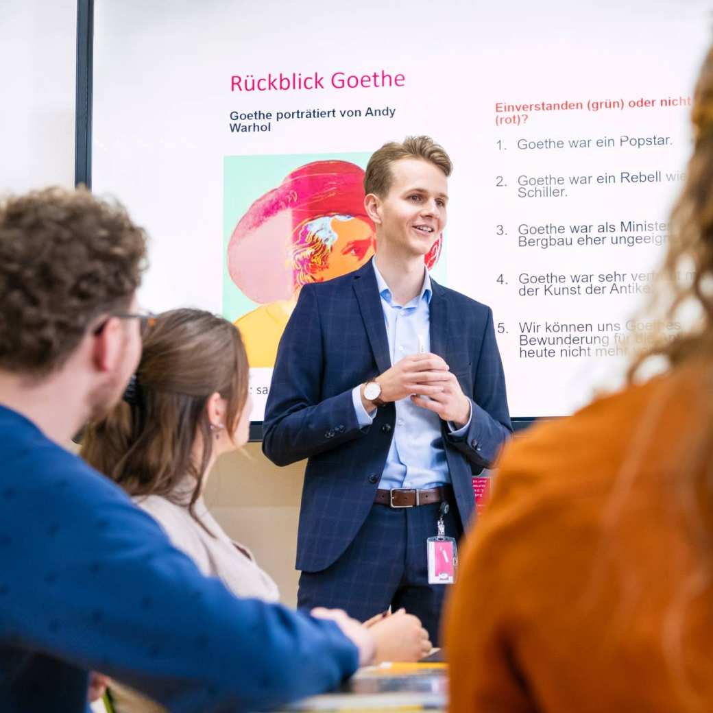 Leraar Duits in pak staat voor de klas met zijn handen in elkaar gevouwen en legt iets uit. Hij heeft een Powerpoint op de achtergrond openstaan.