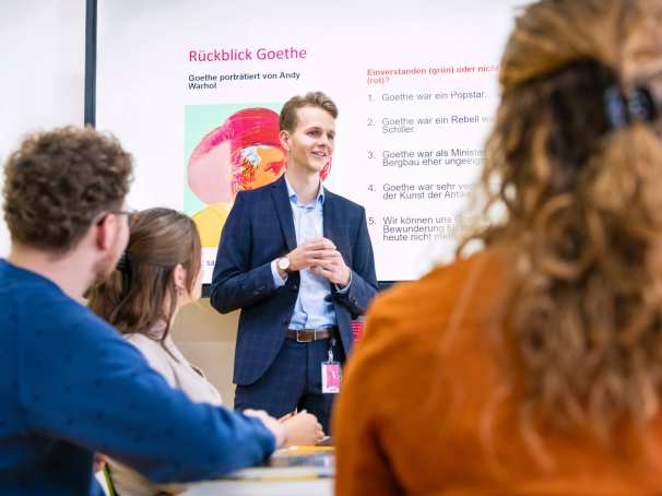 Leraar Duits in pak staat voor de klas met zijn handen in elkaar gevouwen en legt iets uit. Hij heeft een Powerpoint op de achtergrond openstaan.