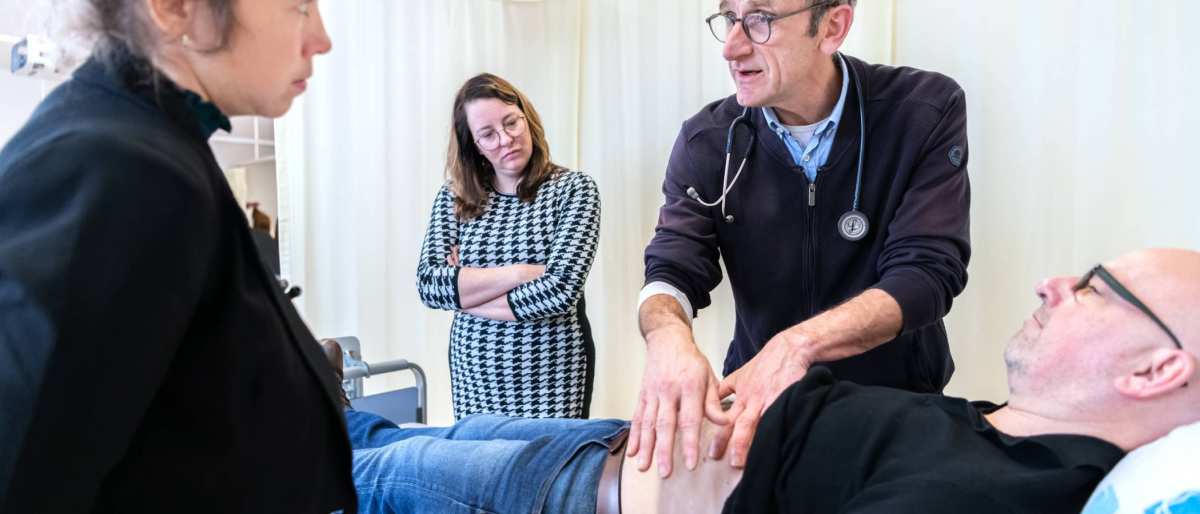 Docent van de HAN Master Physician Assistant geeft uitleg aan 2 studenten over palpatie bij een patiënt op de onderzoeksbank