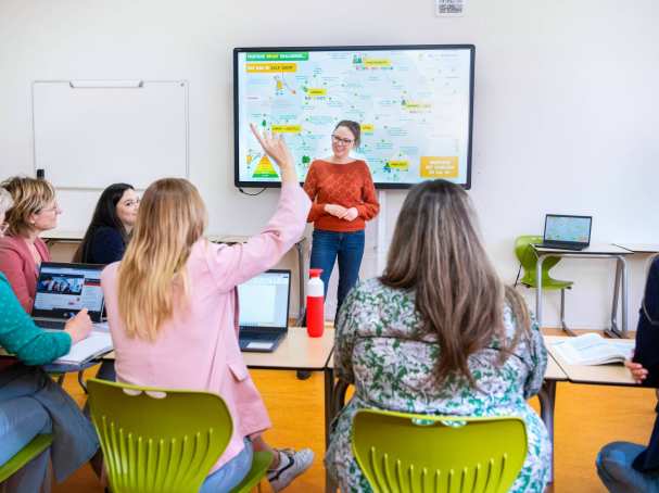 Docent staat voor een volle klas en kijkt lachend naar de student die de vinger opsteekt.