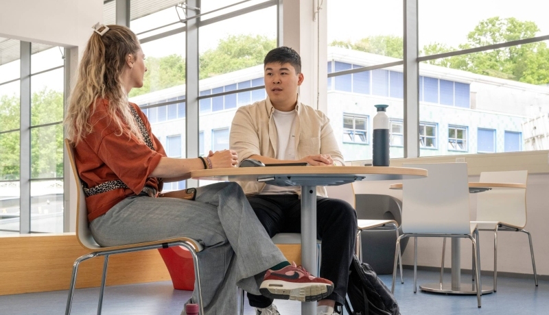 Student in gesprek met docent aan een tafel op podium in hal van gebouw R26, studentbegeleiding. 